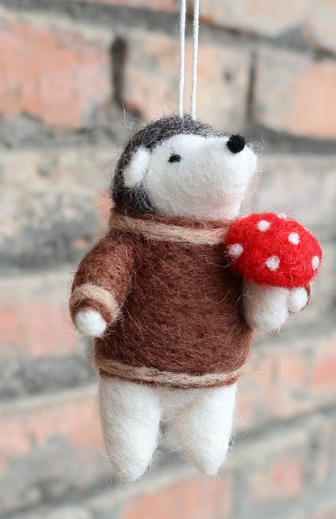A felt hedgehog ornament wearing a brown sweater and holding a red mushroom, Handmade in Nepal.