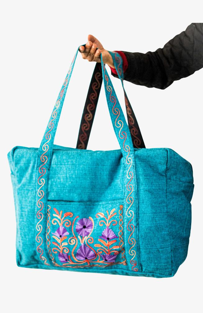 A heavy cotton duffel bag featuring front and back pockets with detailed floral embroidery, handmade in Nepal.