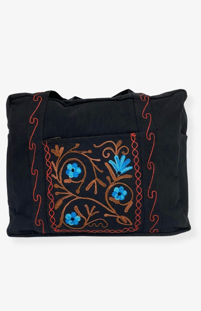 A heavy cotton duffel bag featuring front and back pockets with detailed floral embroidery, handmade in Nepal.