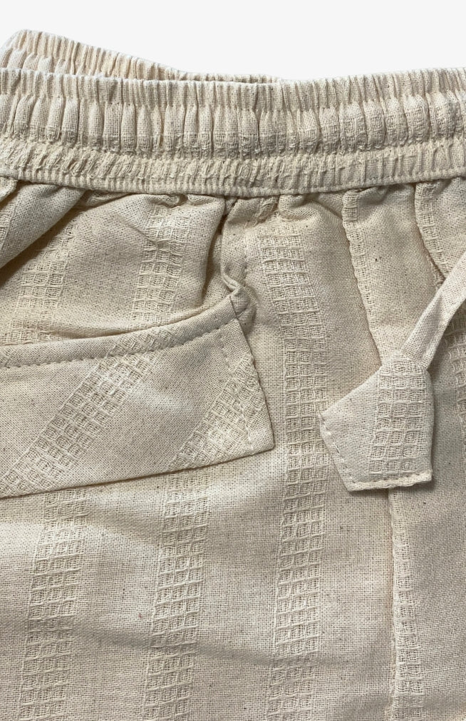 Close-up of a pair of lightweight textured light beige pants showing the elastic waistband, front side pocket, and drawstring detail. Handmade by artisans in Nepal.
