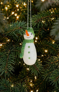 A felt snowman Christmas ornament with a green hat and scarf, orange carrot nose, and green buttons, handmade in Nepal.