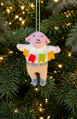 A pig ornament holding colorful flags, wearing a white shirt and beige pants. Handmade in Nepal