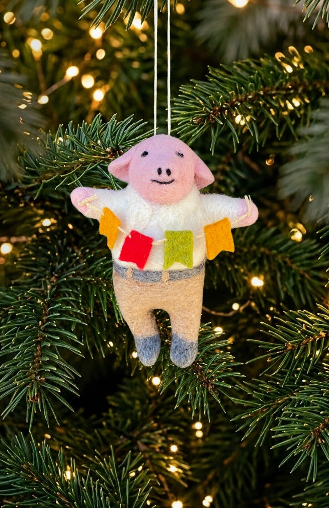 A pig ornament holding colorful flags, wearing a white shirt and beige pants. Handmade in Nepal
