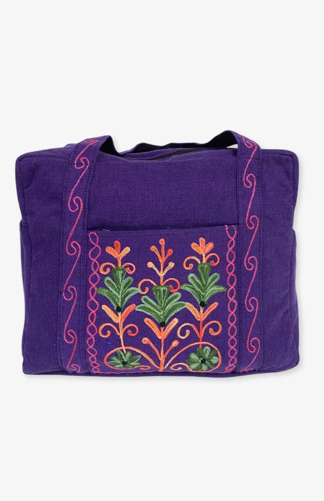 A heavy cotton duffel bag featuring front and back pockets with detailed floral embroidery, handmade in Nepal.