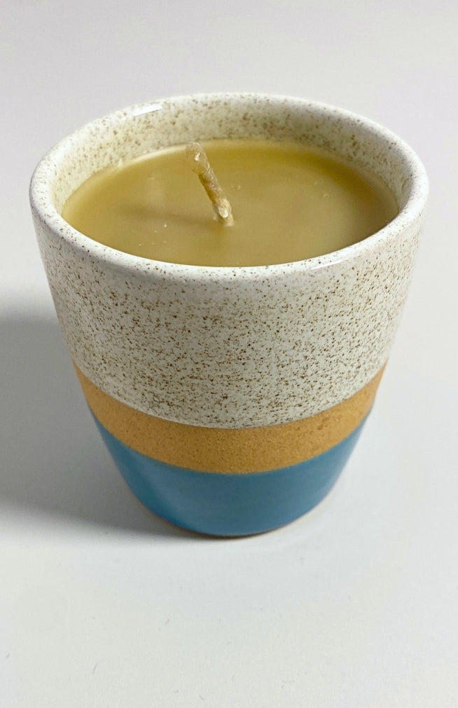A handmade ceramic 100% beeswax candle in a speckled cup-shaped container with a cream upper section, a tan band, and a blue base.