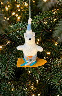 A felted wool polar bear Christmas ornament wearing a blue and white pointed hat and blue swim trunks, riding a surf board. Fair trade Nepal.