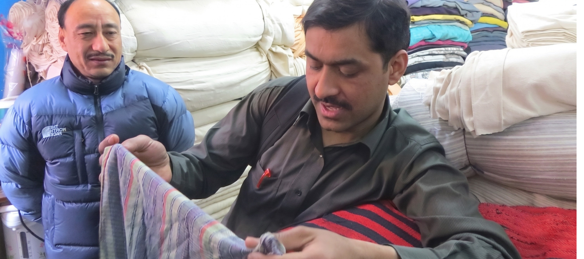 A man in a dark gray shirt holds up a colorful striped fabric sample while another man in a blue puffer jacket observes, set against a background of stacked bedding or fabric rolls.